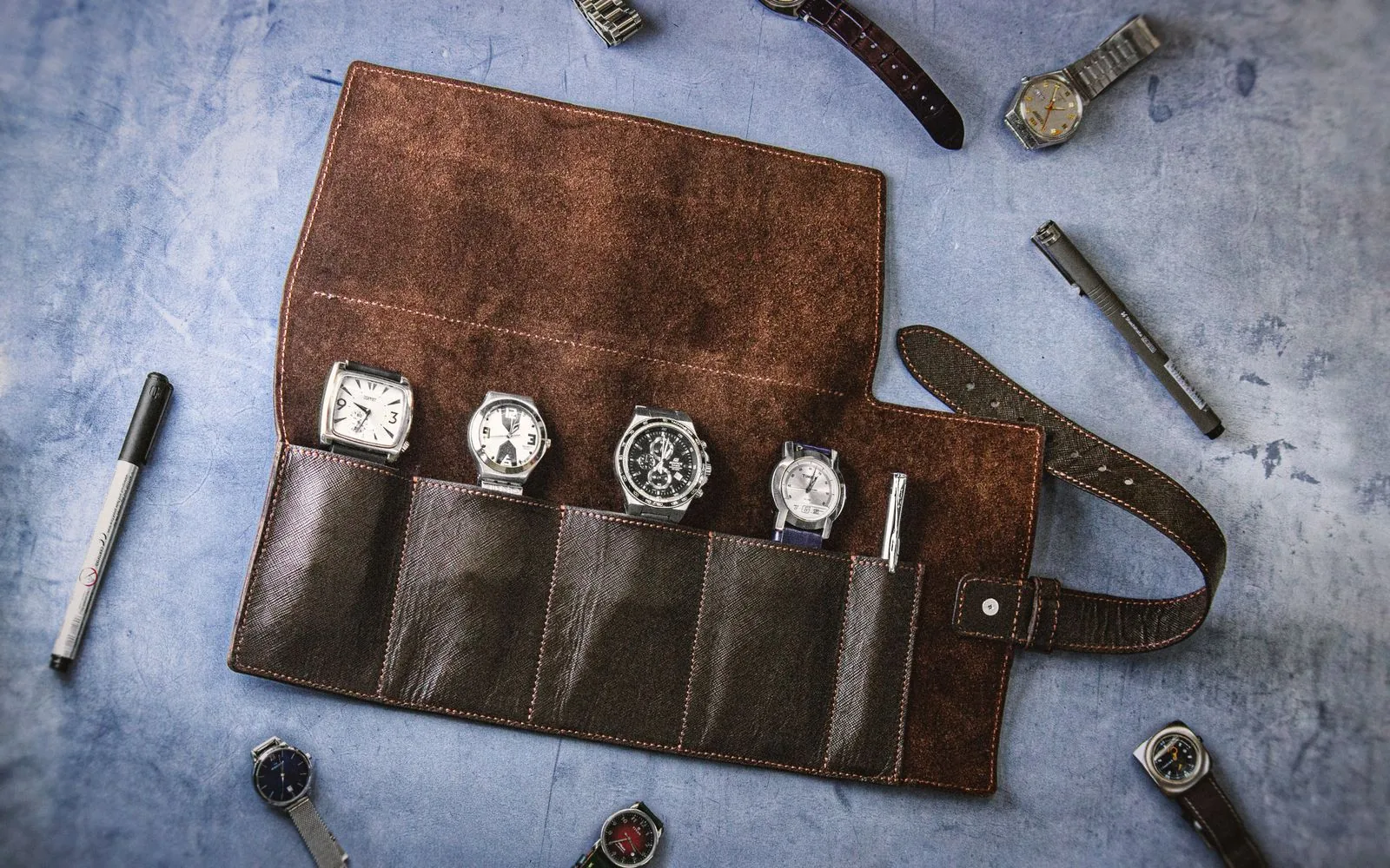 Leather watch roll holding four watches with pens, from above; their own watches guide sizing when buying a watch as a gift