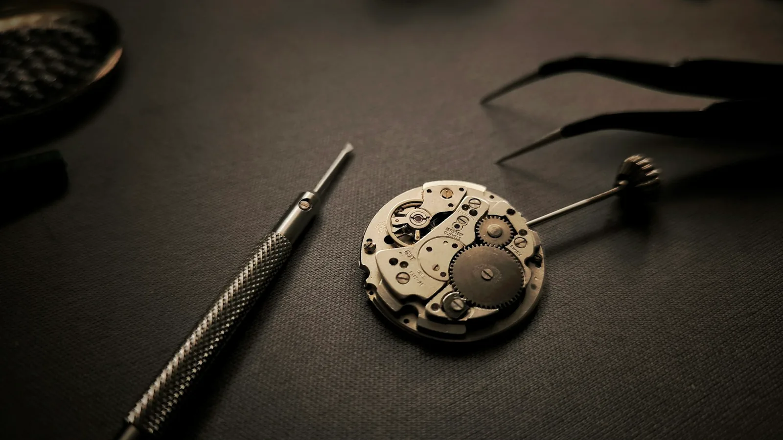 Watch movement, screwdriver and tweezers on a dark bench mat, the kind of work a watch servicing cost covers